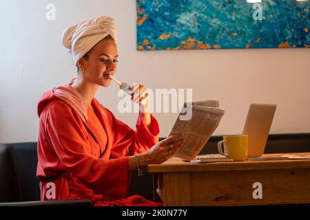 Immagine simbolica Precrastinazione, giovane donna cerca di fare molte cose allo stesso tempo, dopo il bagno, sul computer, telefono, giornale, e colazione Foto Stock