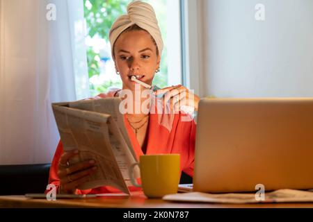 Immagine simbolica Precrastinazione, giovane donna cerca di fare molte cose allo stesso tempo, dopo il bagno, sul computer, telefono, giornale, e colazione Foto Stock