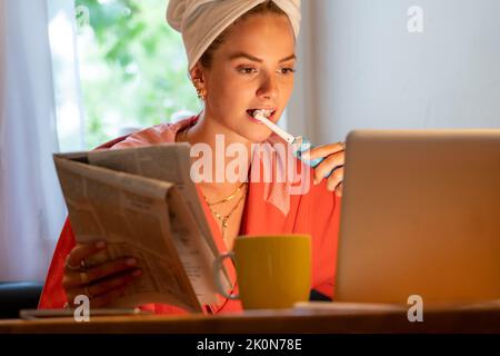 Immagine simbolica Precrastinazione, giovane donna cerca di fare molte cose allo stesso tempo, dopo il bagno, sul computer, telefono, giornale, e colazione Foto Stock