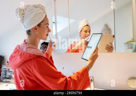 Immagine simbolica Precrastinazione, giovane donna che cerca di fare molte cose contemporaneamente, in bagno, sul computer, parlando al telefono, leggendo le notizie Foto Stock