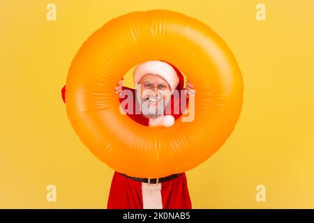 Uomo anziano sorridente con barba grigia in costume di babbo natale con anello di gomma arancione in mano, guardando la macchina fotografica con espressione felice, tour invernale. Studio al coperto isolato su sfondo giallo. Foto Stock