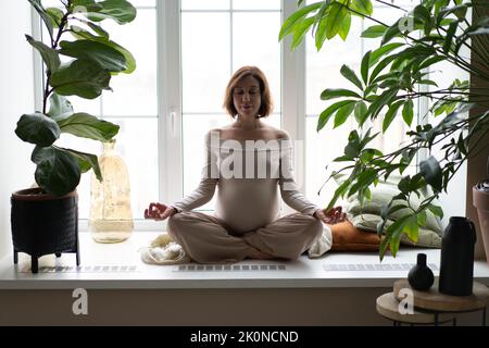 Bella giovane donna incinta sta facendo yoga a casa. Meditazione e relax calmo. Concetto di classi online, quarantena, lezioni online a casa durante una pandemia. Foto Stock