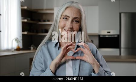 Gentilmente thankful 60s donna anziano volontario maturo femmina pensionato pensionato gry-haired grandy sweetheart rende romantica cornice d'amore da dita forma di cuore Foto Stock