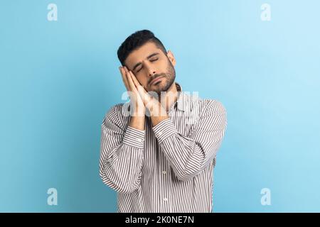 Ritratto di bell'uomo bearded che dorme stendendosi sulle sue palme, avendo sonnellino comodo e riposandosi, dosing fuori, portando la camicia a strisce. Studio in interni isolato su sfondo blu. Foto Stock