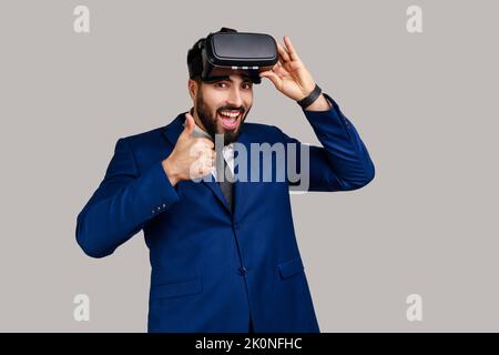 Uomo barbuto con occhiali per realtà virtuale che esprimono felicità dopo aver giocato al simulatore di gioco, mostrando con i pollici in su, indossando tuta di stile ufficiale. Studio in interni isolato su sfondo grigio. Foto Stock