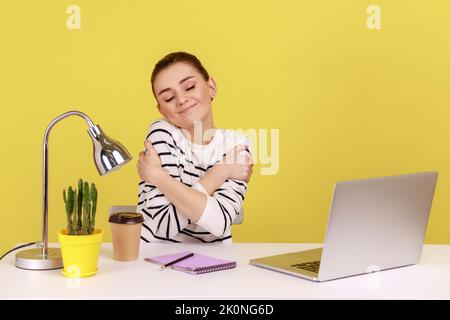 Selfish felice donna ufficio manager abbracciarsi con espressione di piacere, seduta sul posto di lavoro, sentirsi orgogliosi e soddisfatti. Studio interno girato isolato su sfondo giallo. Foto Stock