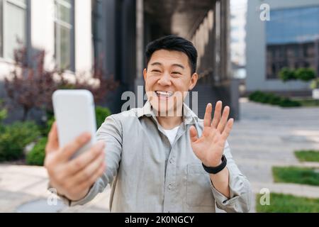 Un uomo coreano che usa lo smartphone per videochiamare, camminare in città fuori, salutarsi al cellulare Foto Stock