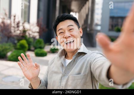 Felice uomo asiatico fare videochiamata tramite cellulare mentre si cammina all'aperto e agitando la mano alla macchina fotografica, gestendo ciao Foto Stock