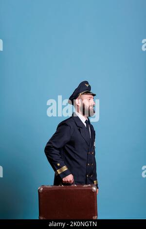 Pilota di aeroplano in uniforme da compagnia aerea professionale che cammina con i bagagli. Aviatore sorridente con bagagli pronti per il volo, a piedi in aeroporto, valigia capitan, vista laterale Foto Stock