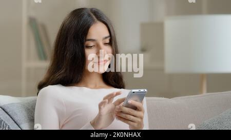 Donna giovane utente freelancer calma attraente ragazza araba donna ispanica si siede sul divano a casa scorre informare le pagine sul telefono dispositivo chatting fa Foto Stock