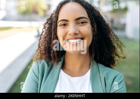 Primo piano di bella positiva sicuro successo ispanica o brasiliana giovane donna con capelli ricci, donna d'affari, in abiti eleganti, in piedi all'aperto, guardando la macchina fotografica, sorride Foto Stock