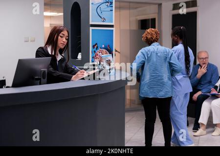 Receptionist femminile che controlla i file di rapporto alla reception dell'ospedale, lavorando sugli appuntamenti di controllo e le consultazioni presso il centro sanitario. Operatore medico che utilizza moduli di assicurazione per aiutare i pazienti. Foto Stock