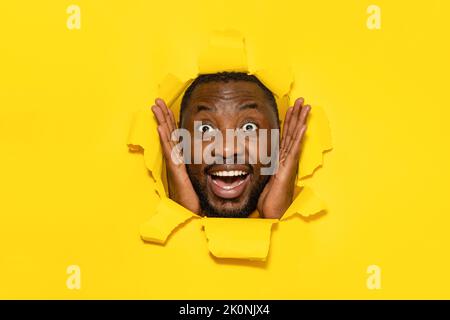 Ritratto di uomo nero sorpreso stupito da offerta commerciale, toccando guance e guardando attraverso il foro in carta strappata Foto Stock