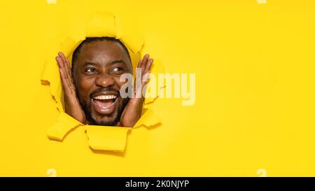 Overjoyed uomo nero stupito da offerta commerciale, guardando da parte lo spazio libero attraverso il foro in carta gialla strappata Foto Stock