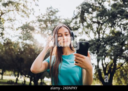 Ragazza adolescente che indossa una t-shirt turchese, con le cuffie, creando playlist sul suo smartphone nel parco. Retroilluminazione del sole Foto Stock