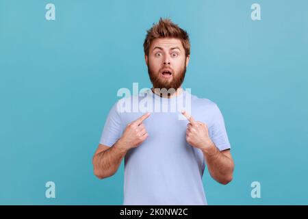 WOW, questo sono io. Ritratto di stupito uomo barbuto fissando in stupore alla macchina fotografica e indicando se stessa, scioccato da successo improvviso, non può credere. Studio in interni isolato su sfondo blu. Foto Stock