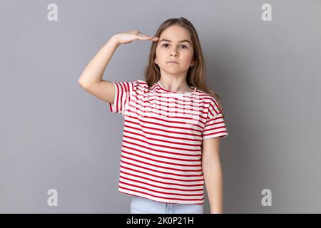 Ritratto di seria bambina con capelli scuri e bossy che indossa una T-shirt a righe in piedi, rendendo saluto con mano sulla testa, accettando l'ordine. Studio in interni isolato su sfondo grigio. Foto Stock