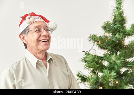 Felice uomo anziano di Latino sorridente accanto ad un albero di Natale che indossa un cappello rosso di Babbo Natale. La felicità delle vacanze. Foto Stock