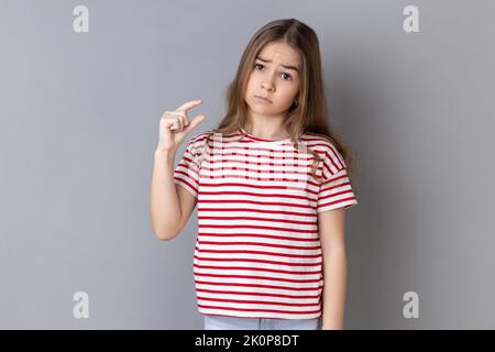 Ne hai bisogno di altro. Bambina che indossa una T-shirt a righe facendo un piccolo gesto e guardando con dispiaciuta implorazione espressione, mostrando minimo. Studio in interni isolato su sfondo grigio. Foto Stock