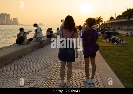 Ragazzi ragazzi e ragazze chiacchierano e camminano durante il tramonto sulla West Kowloon Waterfront Promenade, Hong Kong in serata. Ripresa della luce posteriore Foto Stock