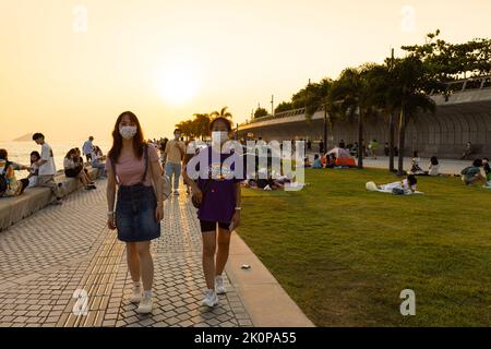 Ragazzi ragazzi e ragazze chiacchierano e camminano durante il tramonto sulla West Kowloon Waterfront Promenade, Hong Kong in serata. Ripresa della luce posteriore Foto Stock