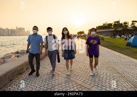 Ragazzi ragazzi e ragazze chiacchierano e camminano durante il tramonto sulla West Kowloon Waterfront Promenade, Hong Kong in serata. Ripresa della luce posteriore Foto Stock