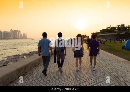 Ragazzi ragazzi e ragazze chiacchierano e camminano durante il tramonto sulla West Kowloon Waterfront Promenade, Hong Kong in serata. Ripresa della luce posteriore Foto Stock