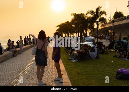 Ragazzi ragazzi e ragazze chiacchierano e camminano durante il tramonto sulla West Kowloon Waterfront Promenade, Hong Kong in serata. Ripresa della luce posteriore Foto Stock