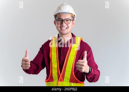 Ritratto dell'ingegnere asiatico che indossa l'hardHat e i pollici in posa contro in uno studio. Architetto, ingegnere, concetto civile di costruzione. Foto Stock