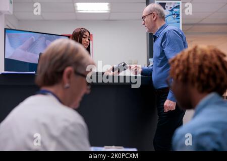 Il paziente utilizza la carta di credito per pagare la visita di controllo e il trattamento con prescrizione medica presso il banco della reception della struttura con l'addetto alla reception. Persona anziana che paga l'appuntamento per un esame nella hall dell'area d'attesa. Foto Stock