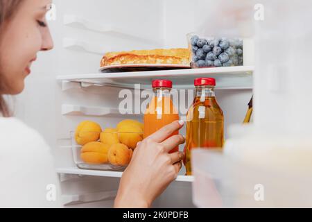 Donna che prende a mano, afferra o raccoglie la bottiglia di vetro del succo dalla mensola aperta del frigorifero o dal cassetto del frigorifero pieno di frutta, verdura, banana, pesche, yogurt. Dieta alimentare sana, concetto di stile di vita Foto Stock