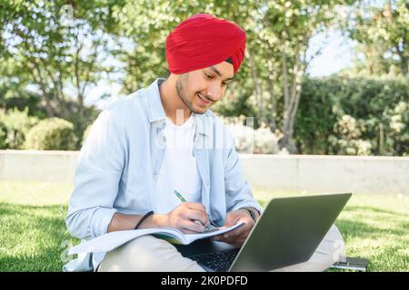 E-learning. Uomo indiano multirazziale in turbante utilizzando il computer portatile per guardare corsi online, webinar, prende appunti seduti nel parco del campus, studente maschile che prepara esami, sostenere corsi di formazione Foto Stock