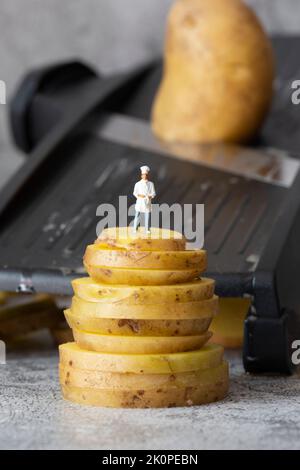 Modello chef in scala ridotta, in piedi su patate impilate, con taglierina da cucina mandolino sullo sfondo Foto Stock