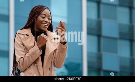 Entusiasta entusiasta sorpresa giovane donna vincitore in piedi all'aperto leggere le buone notizie sul telefono per celebrare la vittoria gioire nella vittoria di successo Foto Stock