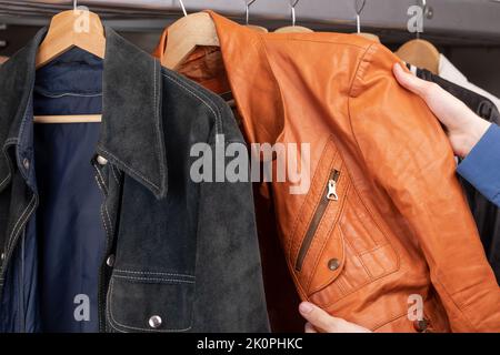 Uomo controlla giacca in pelle arancione in un negozio di seconda mano. Varie giacche d'epoca appendono su una rastrelliera di abbigliamento. Thrifting e sostenibilità in abbigliamento conce Foto Stock