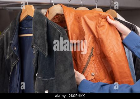 Uomo controlla giacca in pelle arancione in un negozio di seconda mano. Varie giacche d'epoca appendono su una rastrelliera di abbigliamento. Thrifting e sostenibilità in abbigliamento conce Foto Stock