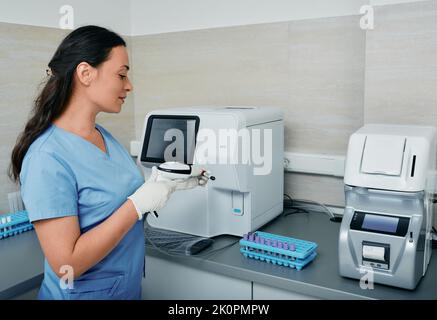 Operatore di laboratorio che esegue la scansione del codice a barre della provetta con sangue sul suo posto di lavoro vicino a una macchina per la ricerca medica Foto Stock