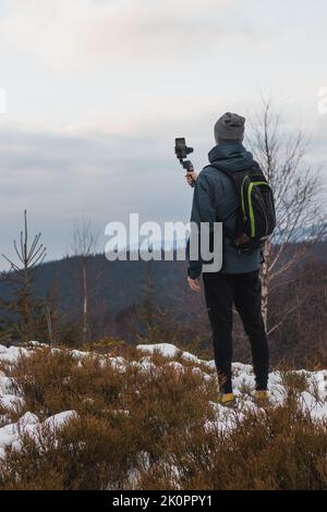 L'uomo in abiti invernali utilizza la tecnologia più recente per vlog e registrare video. Un uomo di mezza età filma su un telefono cellulare attraverso un gimbal. Foto Stock