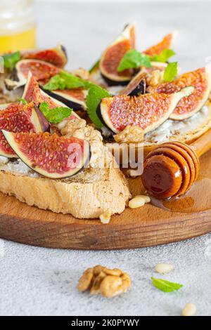 Bruschetta croccante con ricotta morbida, fichi maturi, noci e pinoli, menta e miele su un asse di legno su fondo chiaro. Foto Stock