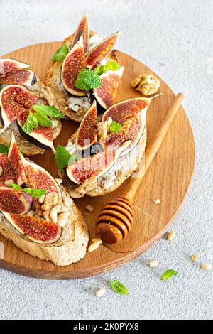 Bruschetta croccante con ricotta morbida, fichi maturi, noci e pinoli, menta e miele su un asse di legno su fondo chiaro. Foto Stock