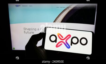 Persona che tiene il cellulare con il logo della società svizzera dell'energia Axpo Holding AG sullo schermo di fronte alla pagina web aziendale. Messa a fuoco sul display del telefono. Foto Stock