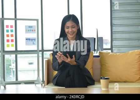 Felice donna asiatica senza busimess che guarda il telefono cellulare per rilassarsi dopo il lavoro in ufficio Foto Stock