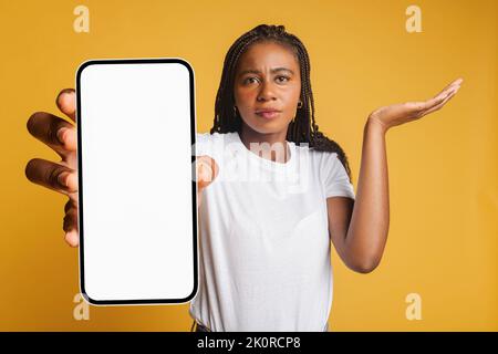 Una giovane afroamericana delusa che tiene in mano uno smartphone e guarda la fotocamera, la donna ha ricevuto una cattiva notifica o messaggi, si sente turbata, frugante, isolata Foto Stock