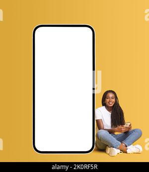 Donna afroamericana seduta vicino a un enorme smartphone con schermo vuoto, con smartphone in mano, chattare nei social network, inviare SMS e sorrisi, scherzare Foto Stock