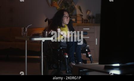Donna con disabilità in una moderna sedia a rotelle motorizzata che entra nel soggiorno e guarda le serie televisive la sera mentre cena Foto Stock