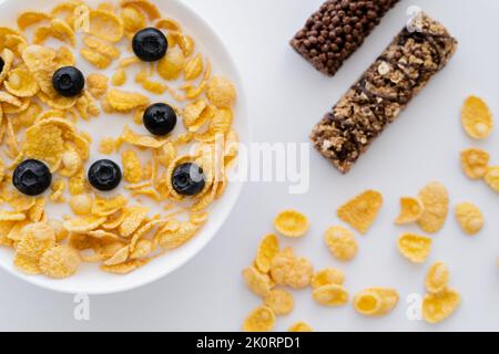 vista dall'alto di corn flakes in ciotola con latte biologico e mirtilli freschi vicino a barrette di muesli isolati su bianco, immagine stock Foto Stock