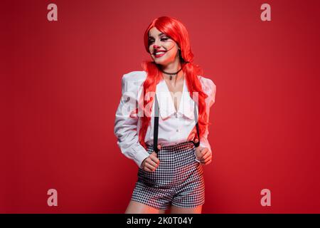 donna redhead con trucco clown posa in blusa bianca e shorts con sospensori isolato su rosso Foto Stock