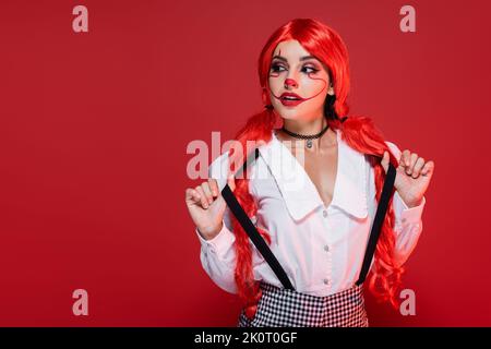 giovane donna rossa in halloween trucco che tocca sospensori e guardando modo isolato sul rosso Foto Stock