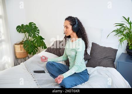 Una giovane donna multietnica siede sul suo letto meditando con l'app di consapevolezza Foto Stock