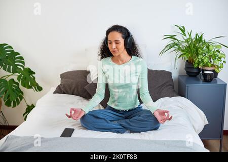 Una giovane donna multietnica siede sul suo letto meditando con l'app di consapevolezza Foto Stock
